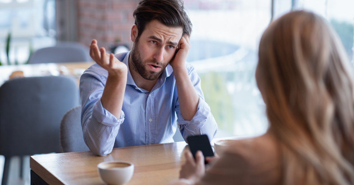 A man sits at a table in a café, gesturing with his hand and looking frustrated whilst talking to a woman on her phone opposite him, is she phubbing him?