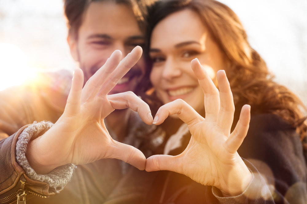 A smiling couple stand close together outdoors, forming a heart shape with their hands showing that relationship counselling is a good idea