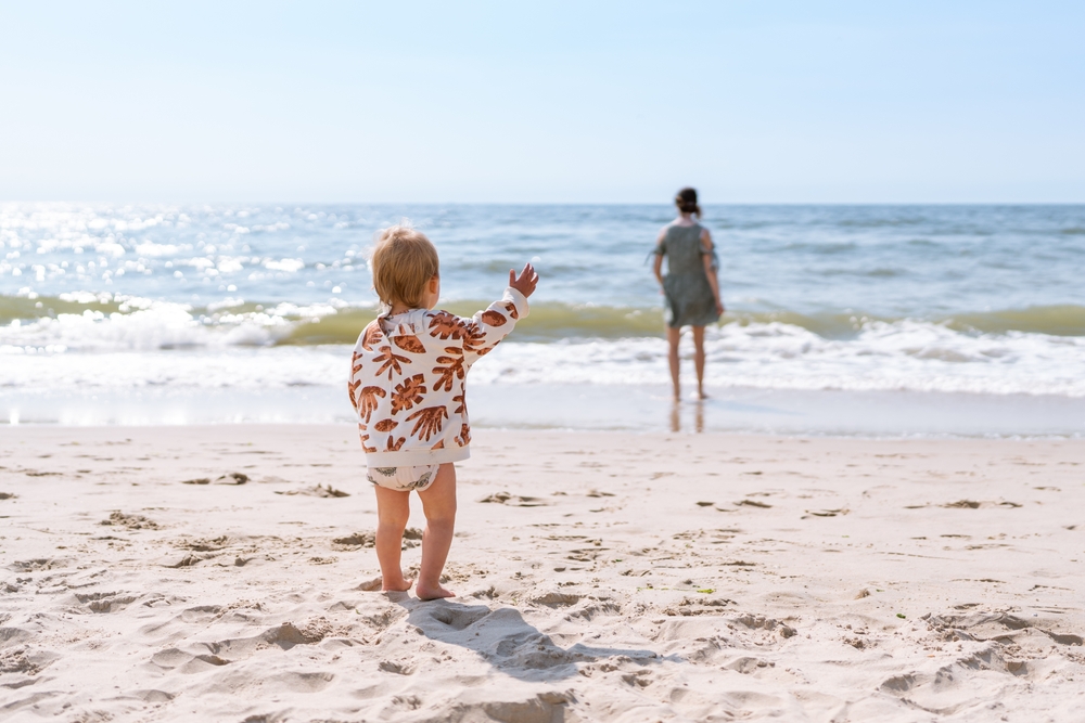 A toddler in a patterned jacket stands on a sandy beach, reaching towards an adult near the shoreline.