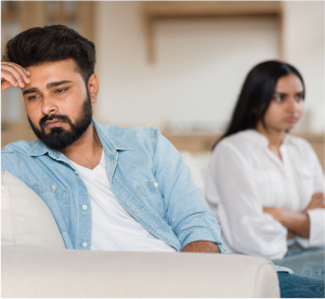 A man sits on a sofa with a troubled expression and a woman sits behind him with her arms folded, can they benefit from online couples therapy?