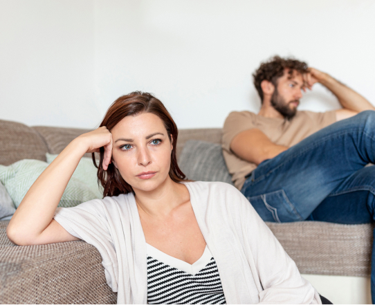 A woman sits at the front of a sofa looking upset, a man sits in the background on the same sofa, looking away with a distant expression.