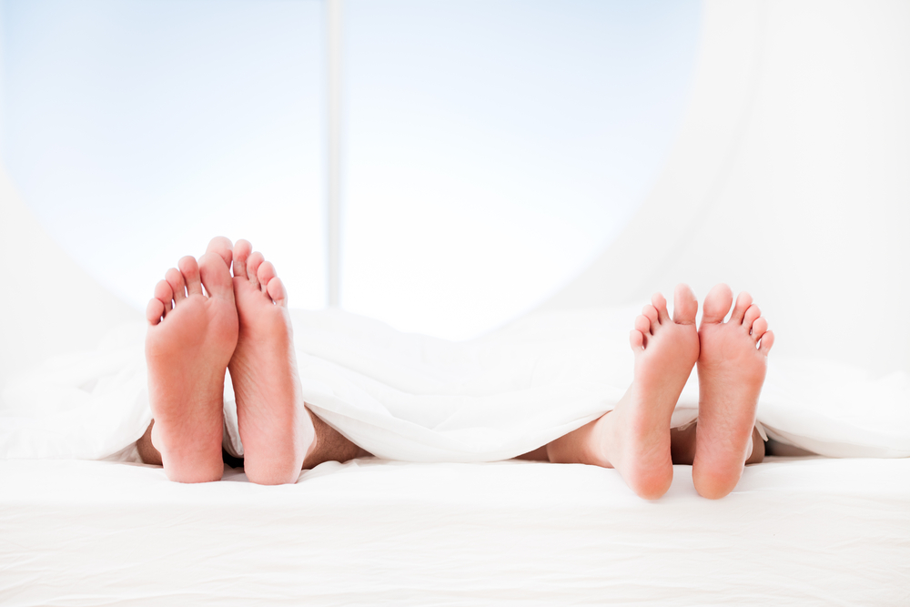 Two people lying side by side in bed with their bare feet sticking out from under a blanket, hinting at intimacy problems between them.