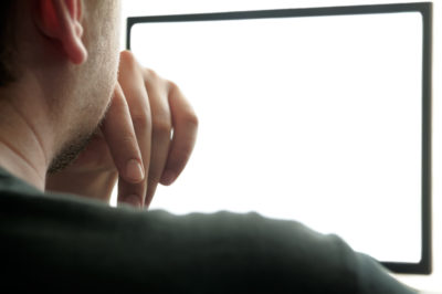 A person seen from behind rests their hand on their chin whilst looking at a blank computer screen, contemplating the impact of porn in relationships.