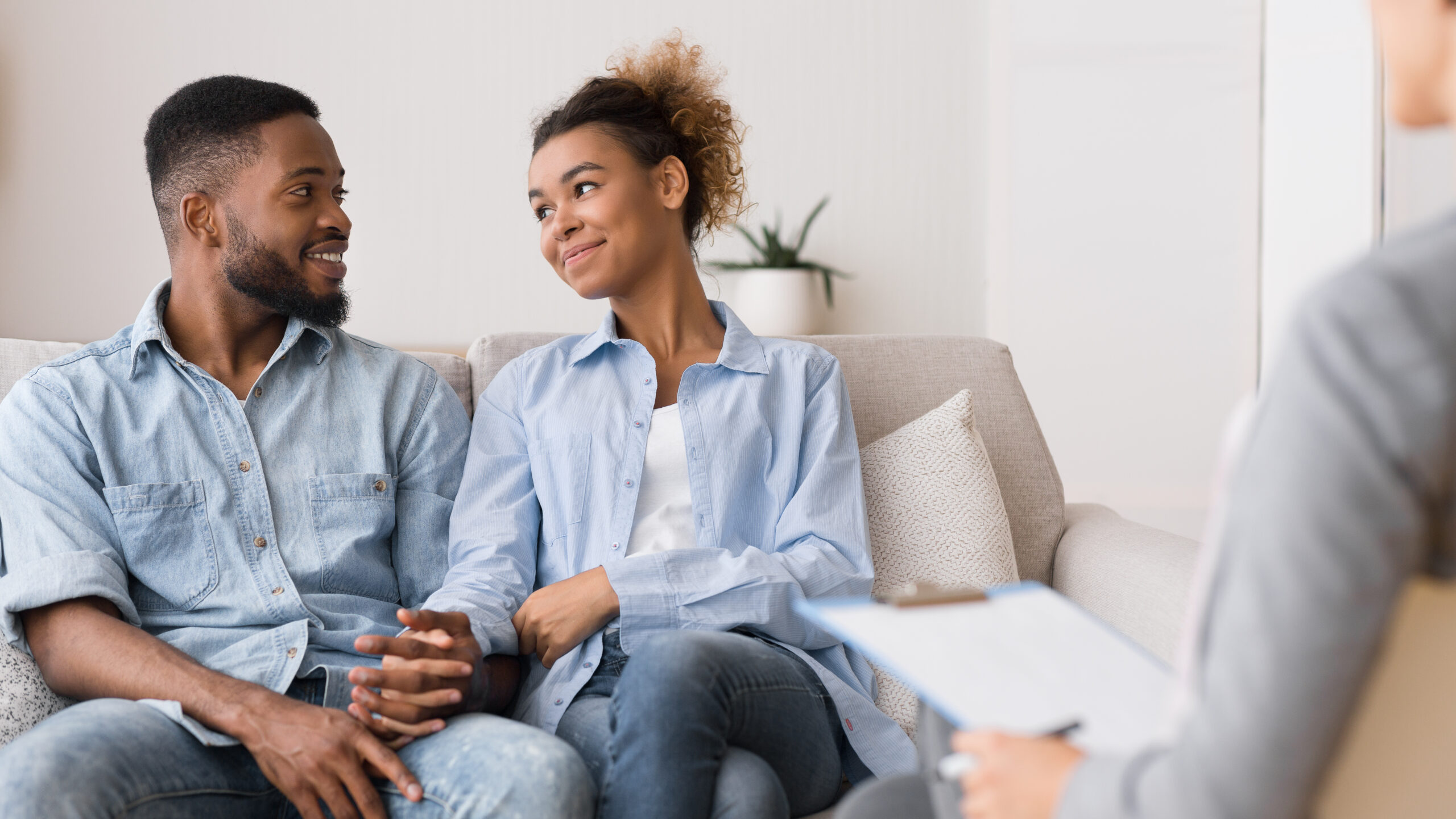A couple sits close together on a sofa, smiling at each other and holding hands, while a person with a clipboard sits nearby, suggesting a relationship counselling session.