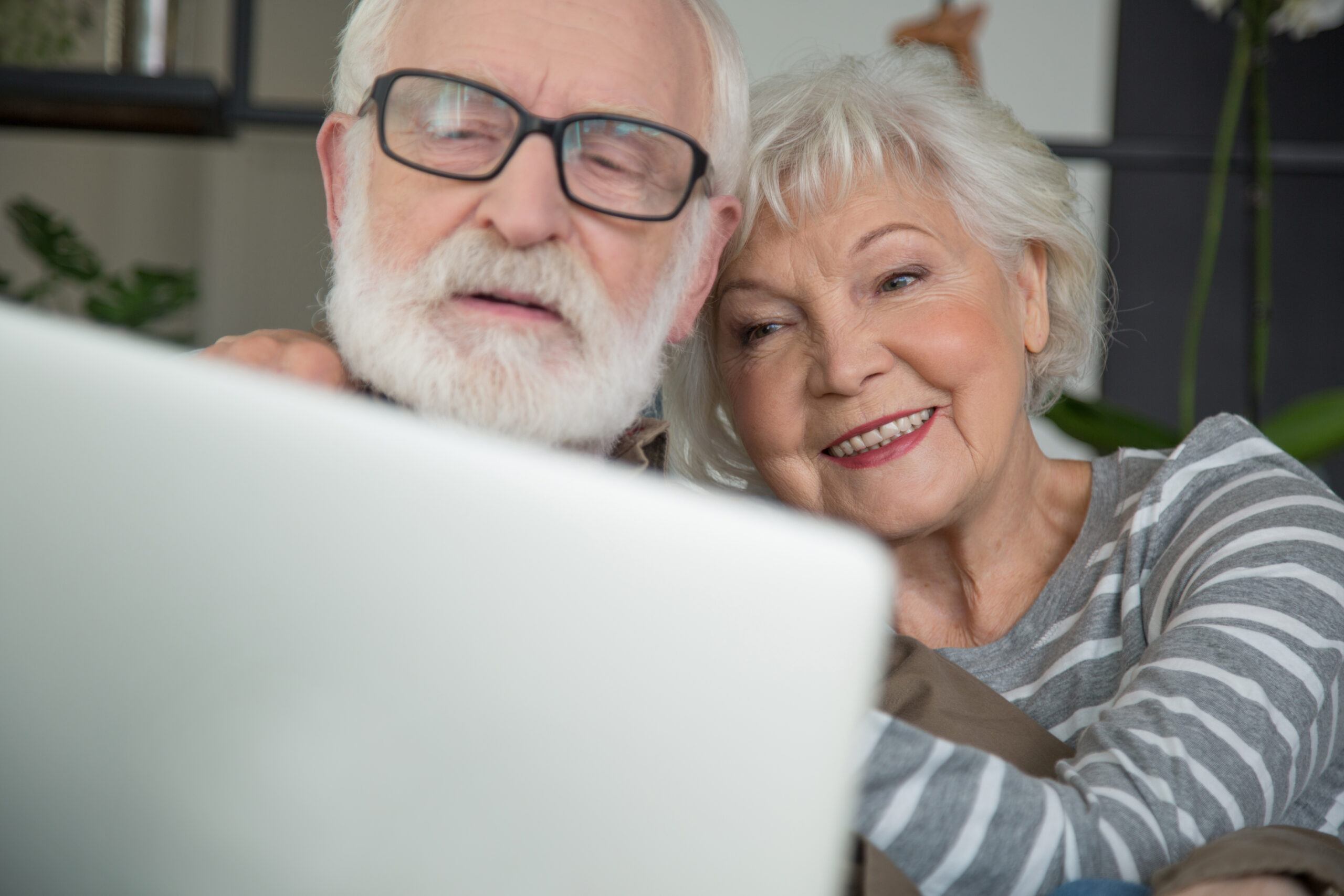 An elderly couple sit close together, smiling as they look at a laptop screen during an online couples therapy session.